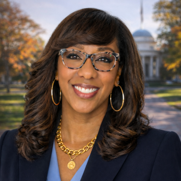 Portrait of a person wearing glasses and a navy blazer smiles in front of campus backdrop.
