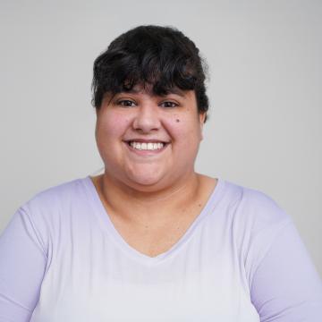A person wearing a light purple top smiles for a portrait in front of a light grey background.