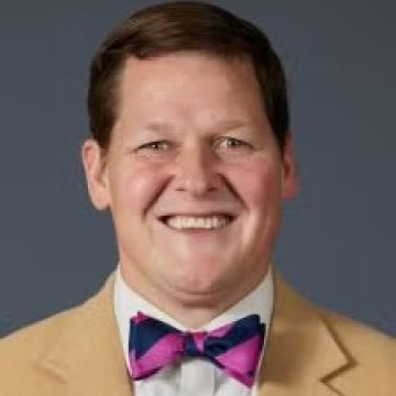 Portrait of man with short brown hair wearing a yellow suit jacket and a pink and navy striped bow tie.