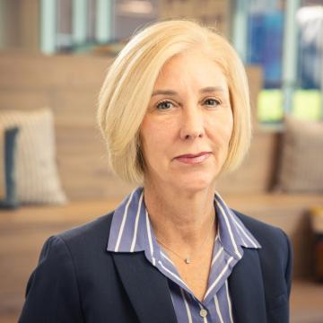 A person with short blonde hair wearing jacket and a striped shirt stand in front of a blurred indoor background.