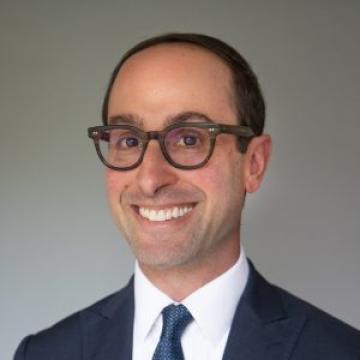 A person with short hair wearing glasses and a tie smiles in front of a gray backdrop.