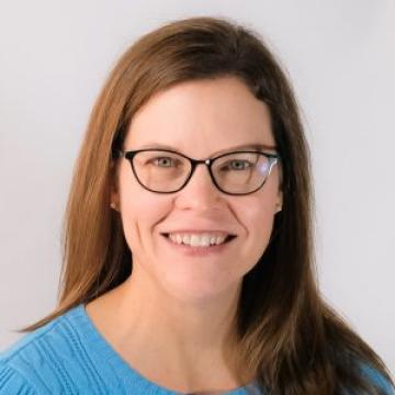 A headshot of a person with brown hair and glasses smiling in front of a white backdrop.