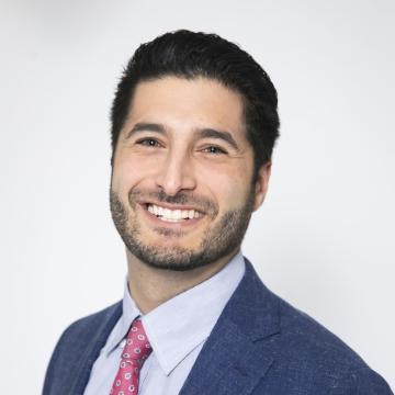 A person wearing a suit and tie smiles in front of a white background.