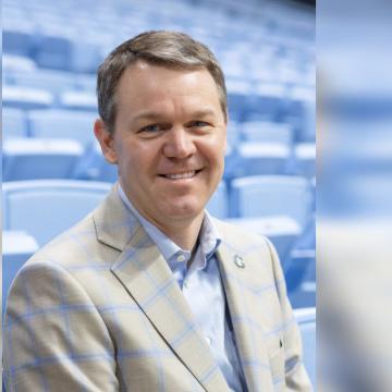 A person smiles wearing a plaid jacket in front of blurred blue stadium seats.