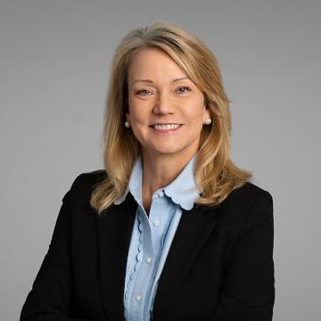 A person with medium length blonde hair wearing a light blue shirt and black jacket smiles at the camera.