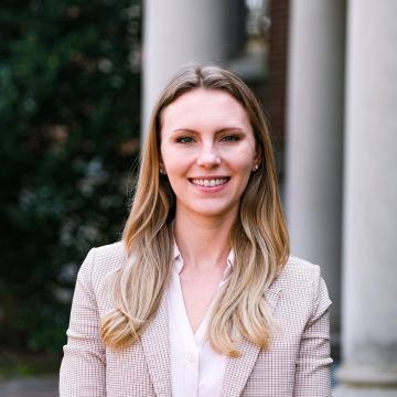 A person with long hair wearing a tan striped jacket stands smiling in front of a building with white columns.