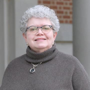 A person with short grey curly hair and classes stand smiling outdoors in front of a brick building.