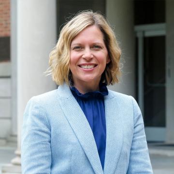 A portrait of a person smiling with short blonde hair wearing a light blue jacket standing in front of a building column.