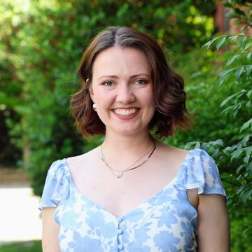 Person with short brown hair smiles at the camera standing in front of green bushes outside.