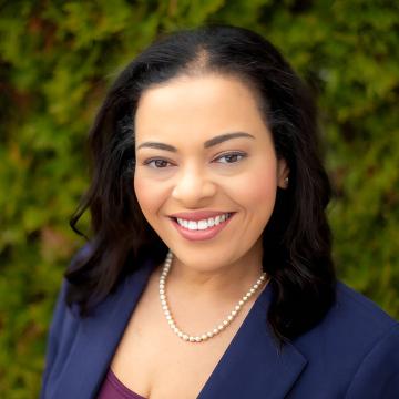 A person with black hair wearing a navy jacket smiles standing in front of a blurred green background.