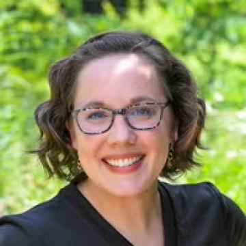 Staff portrait of a smiling person with short wavy hair wearing glasses against a blurred green background.