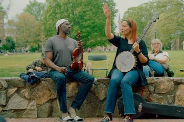 Two musicians sit on a stone wall, one holds a violin and one holds a banjo. 