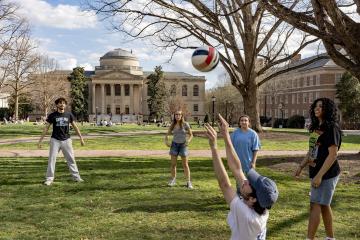 Students stand in a circle passing a volleyball in a grassy area surrounded by trees and buildings.