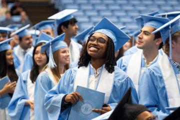 A student wearing a light blue cap and gown holding a diploma smiles with other graduates visible in the background.