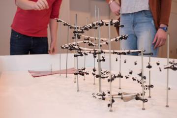A person holds a small ball above a curved track set up on a table top, about to release the ball so that it can roll down