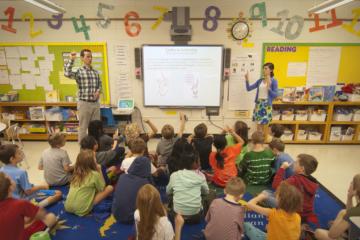 Teachers standing at the front of a classroom with students seated on the carpet below