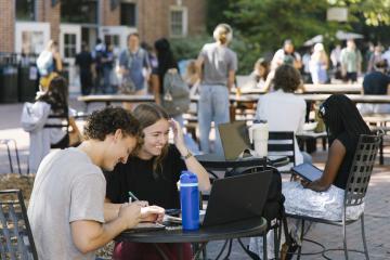 Students sit at outside tables on campus and complete work. 