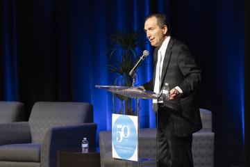 Person in suit and tie stands at podium and speaks into microphone.