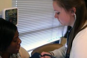 A woman wearing a stethoscope wraps a blood pressure cuff around a seated woman's arm. 