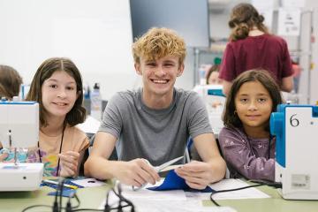 Austin Roebuck and two ArtsCenter summer campers smile at the camera while working with sewing machines and fabric in a classroom.
