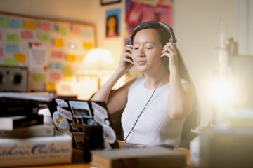 Person holds headphones up to ears while peering down at laptop.
