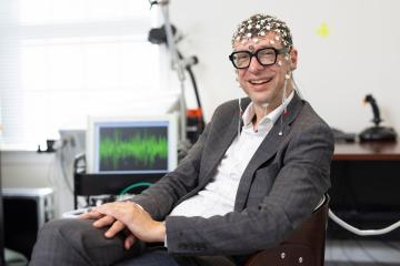 Professor Flavio Frohlich faces the camera seated at a desk wearing tACS head-gear