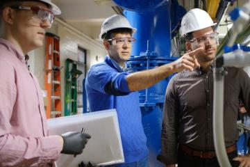 Three men in hardhats stand in a water treatment plant. The man in the middle is pointing at a piece of equipment.