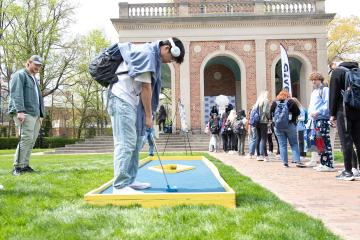 A UNC student wearing headphones plays miniature golf with students lined up at the Carolina Bell Tower in the background.