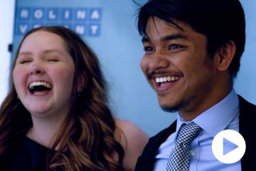 Two smiling Carolina Covenant Scholars with a blue Carolina Covenant sign in the background and a video play button in the bottom right-hand corner.