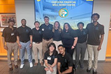 A group of Carolina students pose for a portrait in front of a screen with the words Carolina Financial Well-Being Center written on it.