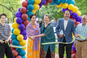 Five people smile for the camera at a ribbon-cutting ceremony under a balloon arch.