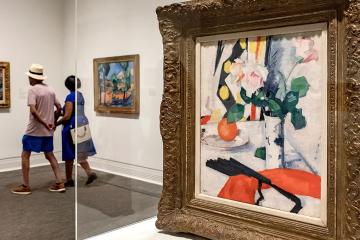 Painting sits in a glass box on display with person passing in the background.