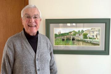 Bob Lauterborn smiles while standing next to a painting by his late wife on the wall in the background