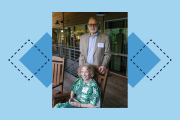 An older man stands behind a seated woman on a wooden patio, both smiling, with glass doors and a railing in the background.