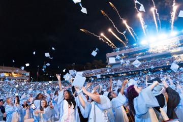 Carolina students throw caps into the air with fireworks in the background during commencement at Kenan Memorial Stadium.