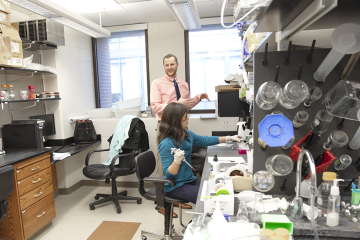 Chad Pecot stands speaking in lab, while a woman works at a workbench.
