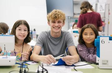 Austin Roebuck and two ArtsCenter summer campers smile at the camera while working with sewing machines and fabric in a classroom.