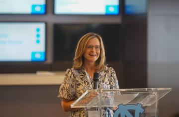 Woman with short blonde hair and glasses smiles while speaking at a podium.