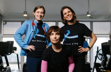 Eli Levene sits and looks at the camera with Gabby Brennan and Carly Bailey standing and smiling on either side of her.