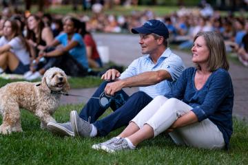 Kevin and Amy Guskiewicz sit on a lawn, with Kevin holding his dog Charlie on a leash.