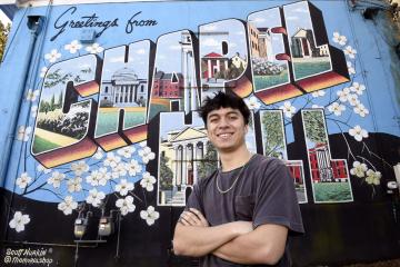 gustin Orozco smiles in front of a mural that reads 'Greetings from Chapel Hill.