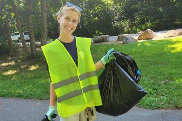 Eloise MacLean faces the camera outdoors wearing a yellow vest and holding a trash bag