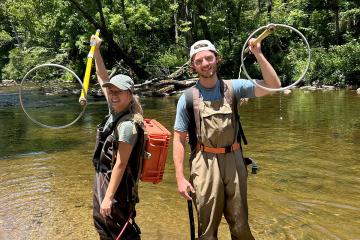 Two people stand side by side in a river wearing electrofishing equipment