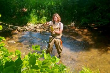 Tayton Alvis stands in a river holding a clipboard, taking notes