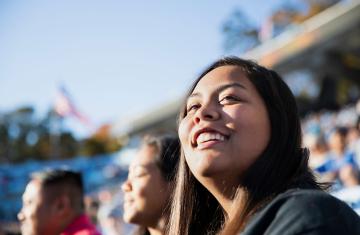 A smiling student sits in the stands at a football game looking out into the horizon