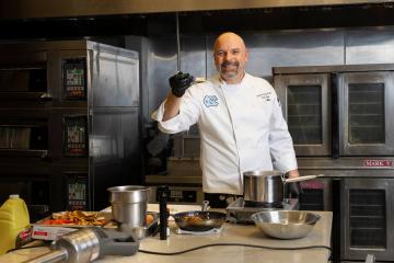Christian Chiron standing in a kitchen in a chef&rsquo;s coat while smiling and holding up ingredients on a spoon.