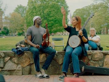 Rhiannon Giddens sitting on a stone wall on Polk Place while holding a banjo, next to a musician holding a fiddle.