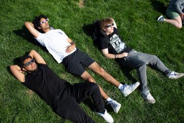 Three students lie down on the grass with glasses on and peer up at the sun.
