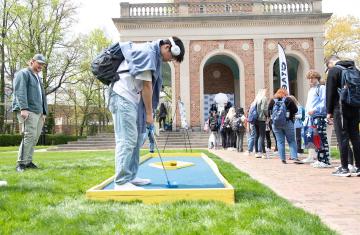 A UNC student wearing headphones plays miniature golf with students lined up at the Carolina Bell Tower in the background.