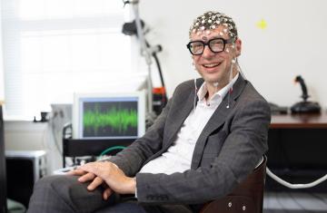 Professor Flavio Frohlich faces the camera seated at a desk wearing tACS head-gear